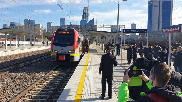 Widok na stację Warszawa Główna, podróżni stojący na peronie i pociąg stojący przy peronie.