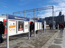 Peron z tablicą z nazwą stacji Warszawa Główna, w tle podróżni.